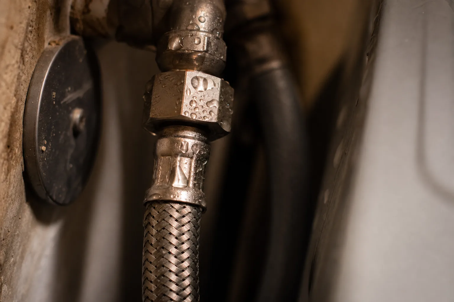 A braided stainless steel washing machine supply line coupling behind an appliance, water beaded on the metal