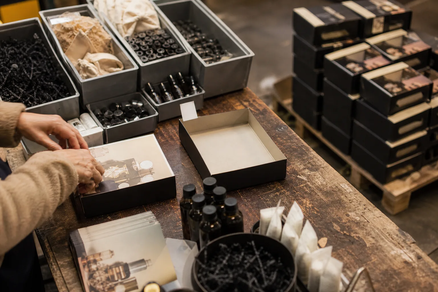 A warehouse workbench where a worker assembles retail gift sets, with component bins, printed inserts, empty branded boxes, and finished kits stacked on a pa...