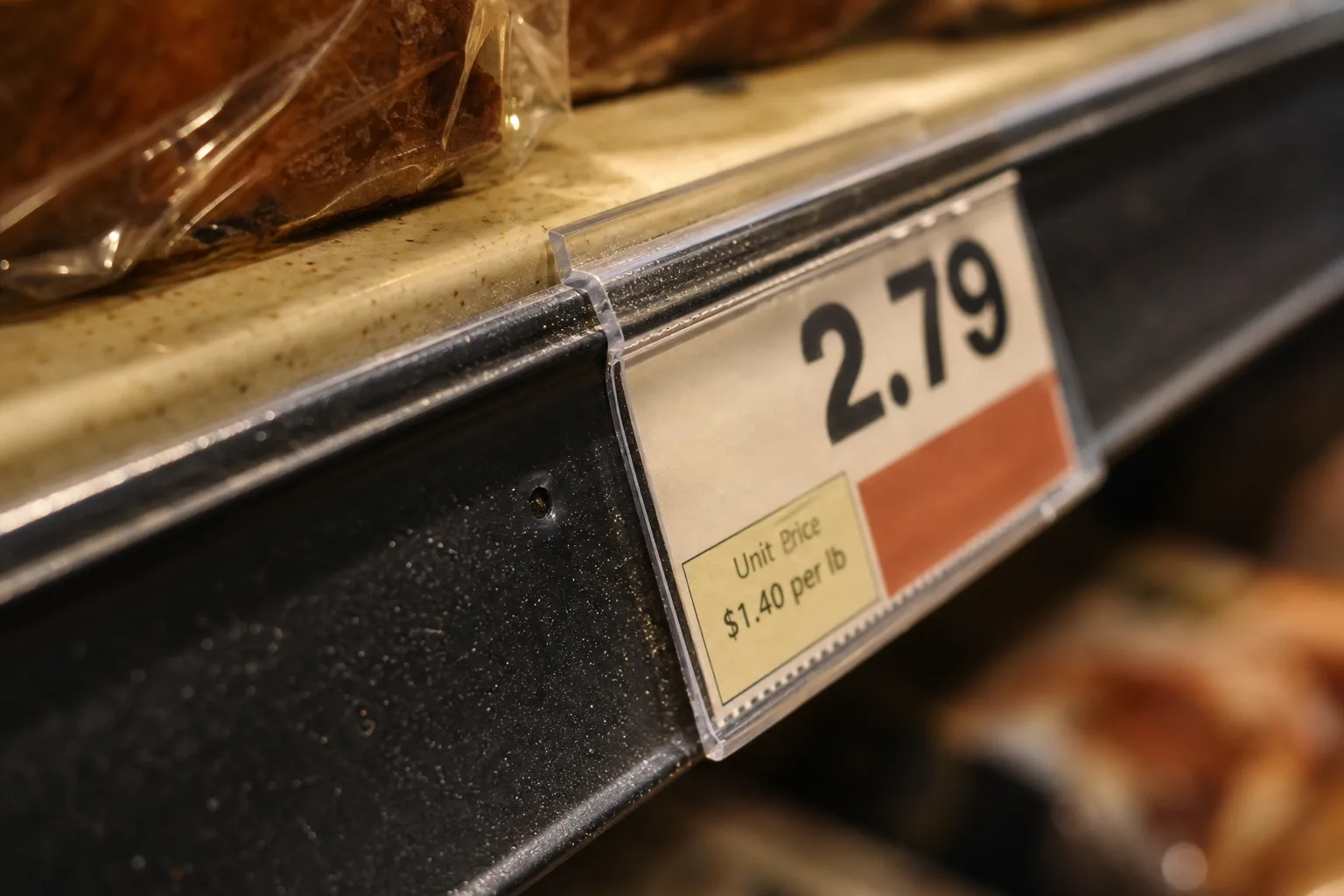 A supermarket shelf edge with a price tag clipped to the rail, the small unit price text catching the light
